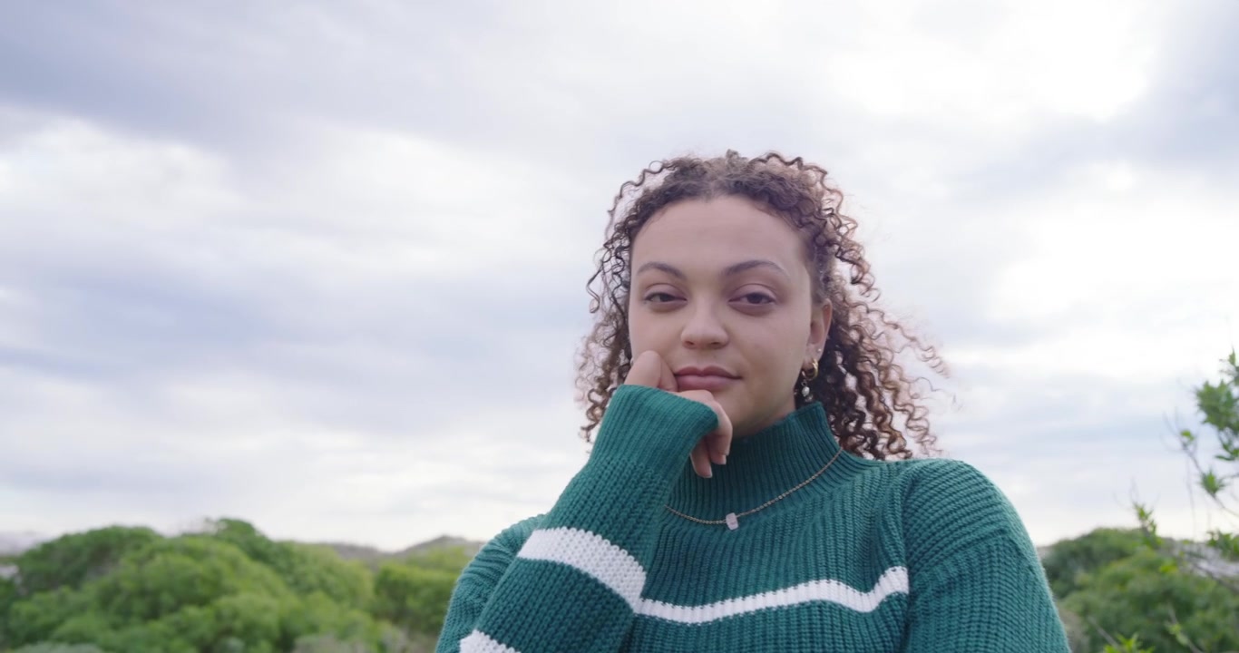 Enfoque de cámara en una mujer afroamericana adulta, descruzándose, sonriendo en las dunas y vistiendo un suéter de punto verde.