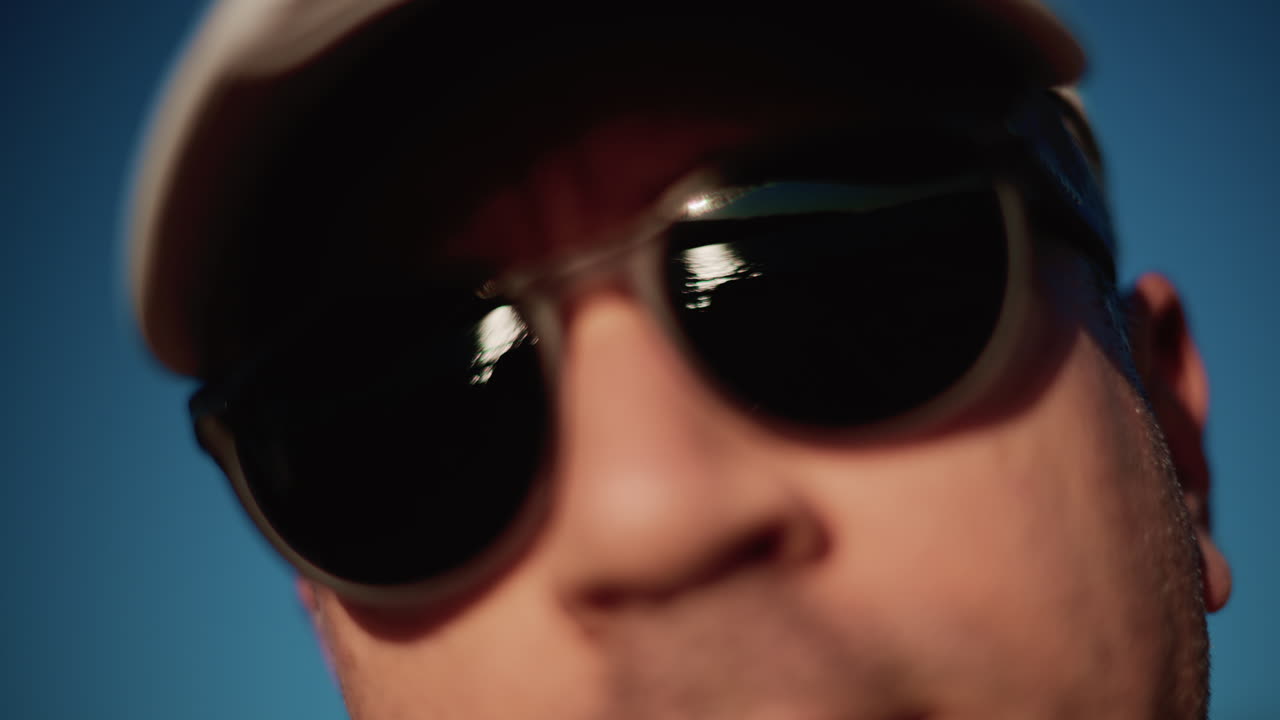 Extreme close up of a man wearing dark sunglasses and a cap, framed tightly against a clear blue sky in strong daylight
