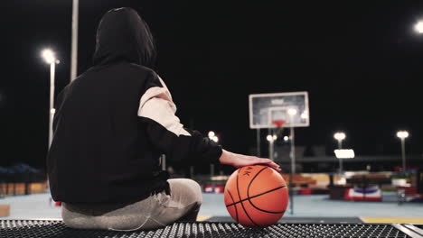 Rückansicht-Einer-Basketballspielerin-Im-Hoodie,-Die-Mit-Ball-Sitzt-Und-Nach-Ihrer-Trainingseinheit-Nachts-Auf-Dem-Außenplatz-Eine-Pause-Macht