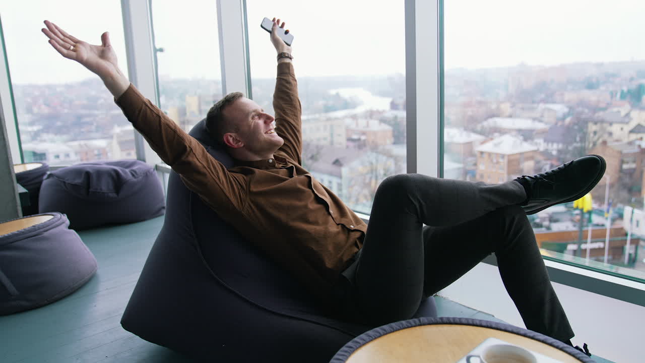 Relaxed businessman indoors. Happy young man is sitting by the window with stretched arms and having a rest in office on the city view window background.