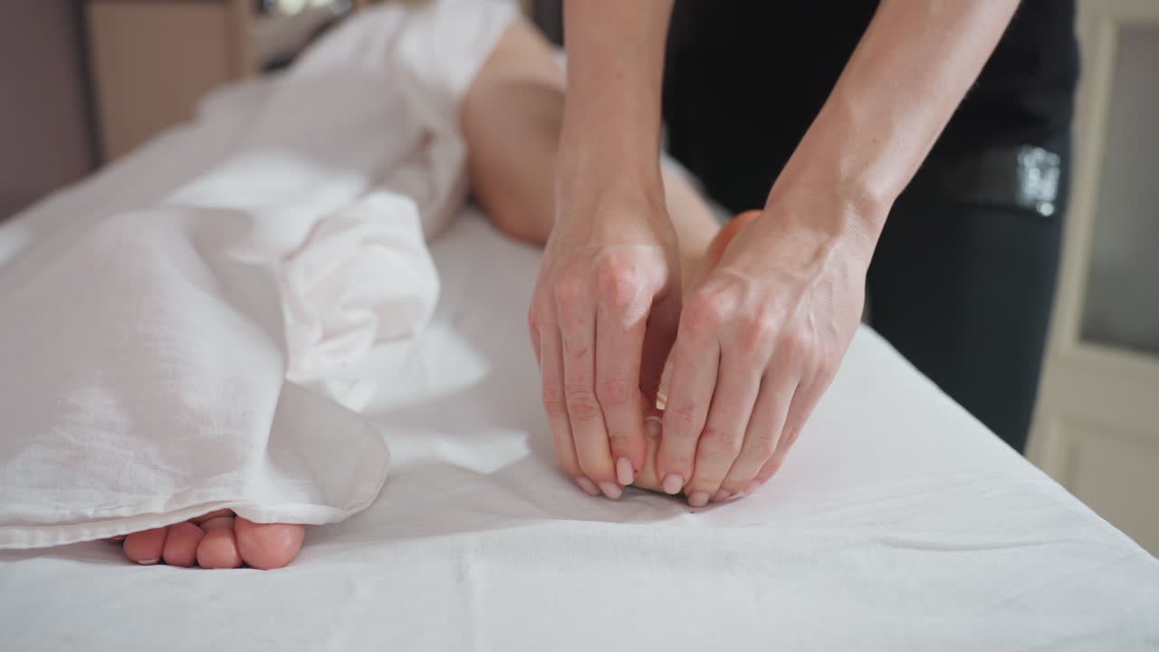 Massage therapist gently pressing client back foot with both thumbs while folding toes during professional wellness session, showing focused hand technique on clean surface with soft natural light