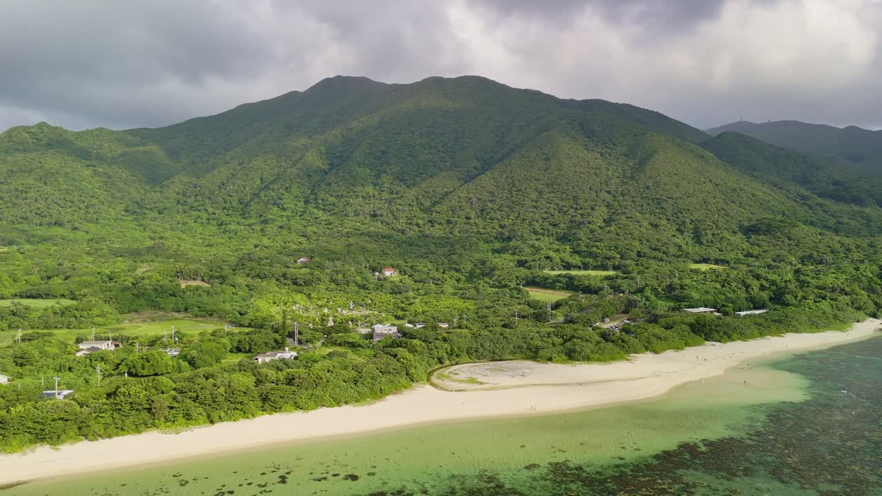 Japonya'nın Okinawa bölgesindeki Ishigaki Adası'nın yükselen, yemyeşil dağ zirvelerine karşı konumlanmış Sukuji Plajı'nın beyaz kumlarını ve zümrüt mercan resiflerini gösteren sinematik drone çekimi