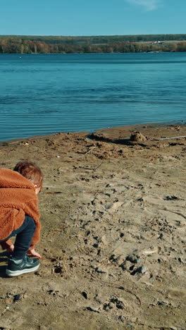 Caucasian boy touching sand on the river bank. Cute kid takes some handful of sand and throws it into water. Vertical video