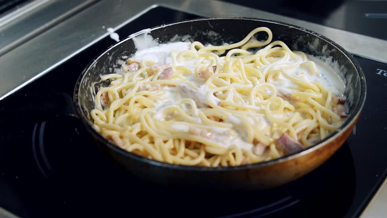 Process of cooking tasty pasta. Chef is pouring white sauce into spaghetti cooking in a frying pan. Preparing creamy pasta. Close-up