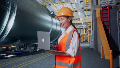 vista lateral de una ingeniera asiática con casco de seguridad trabajando en una computadora portátil mientras está de pie en una fábrica de fabricación de tuberías