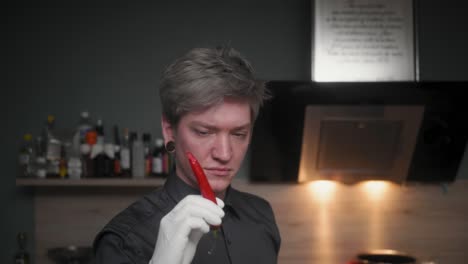 attractive professional male chef in an elegant black shirt with an alternative look biting off a red chili, looking into camera a modern, fancy looking kitchen, slow motion