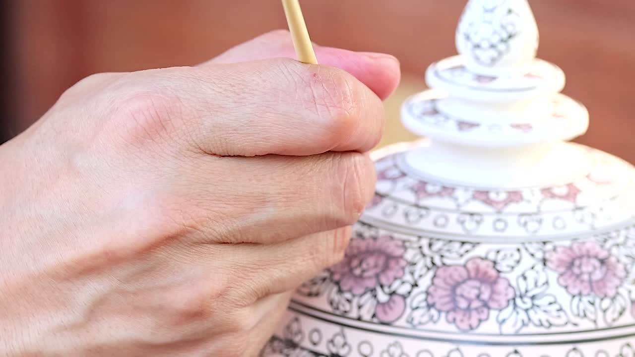 A detailed view of a hand painting intricate floral designs on a ceramic lid using a fine brush.