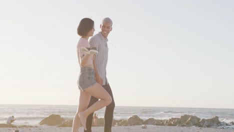 Happy-biracial-couple-walking-and-holding-hands-at-beach,-in-slow-motion