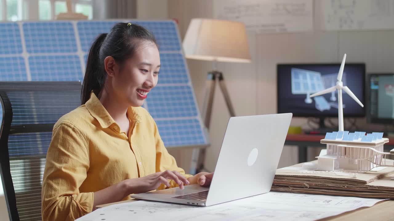 사무실에서 태양 전지판과 풍력 터빈을 가진 작은 집의 모델 에 노트북에서 타이핑하는 미소 아시아 여성의 측면 시각