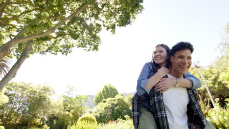 happy diverse couple playing piggyback and walking in sunny garden, copy space, in slow motion