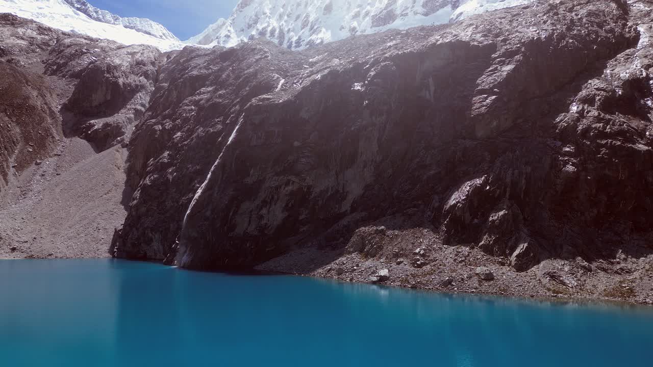 Low flight across vivid blue mountain lake toward cliff below mtn peak
