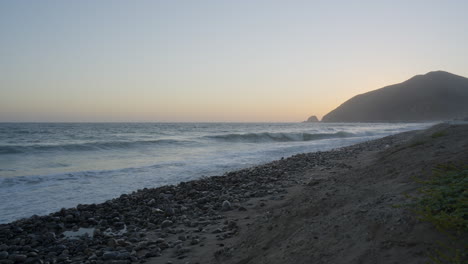 Toma-Estacionaria-De-Olas-Rodando-Por-El-Océano-Pacífico-Con-La-Puesta-De-Sol-Detrás-De-Una-Montaña-Distante-Ubicada-En-El-Sur-De-California