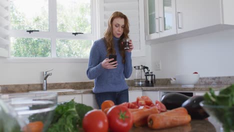 mujer bebiendo café mientras usa un teléfono inteligente en la cocina