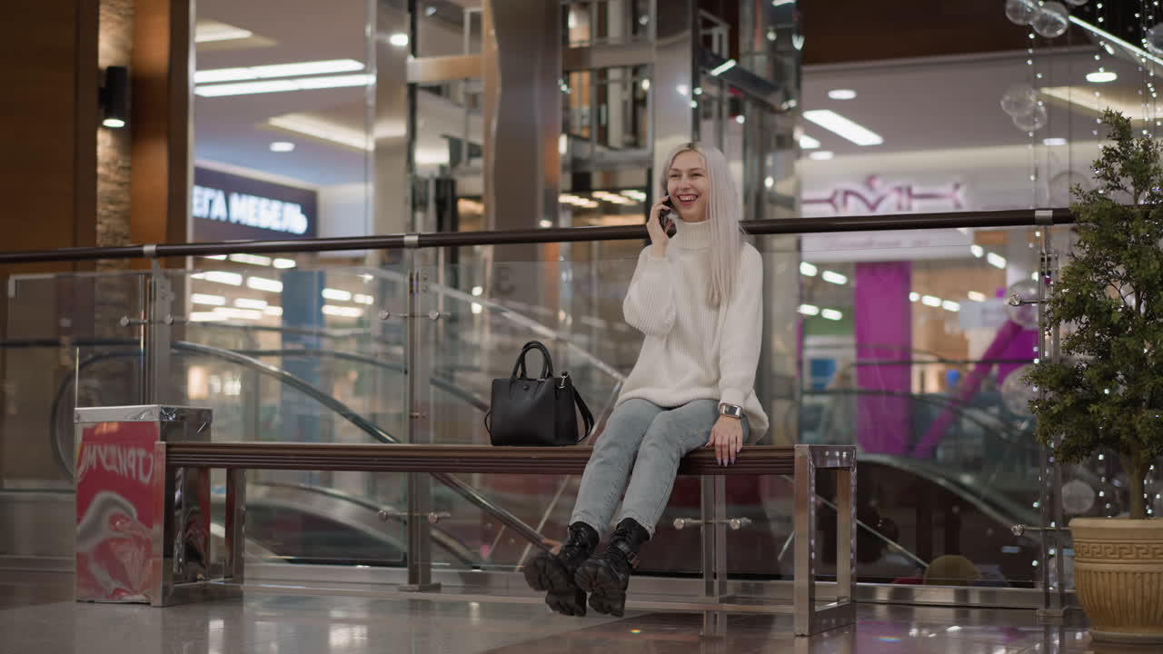 beautiful young lady seated on bench giggling into smartphone call, playfully lifting leg in knitted sweater, denim and chunky black boots in modern mall interior with moving elevator behind
