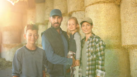 portrait of happy caucasian family of farmers with little girl and teen boy standing at stable with hay stocks and smiling at camera