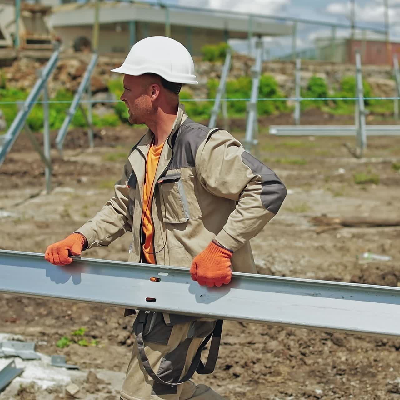 Construction site of a solar farm outdoors. Worker in safety helmet carries metal frame on the field.