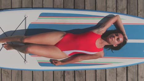 Aerial-View-Of-A-Girl-In-Red-Swimsuit-Lying-On-The-Surfboard-With-A-Paddle-Next-To-Her-On-The-Wooden-Harbor-1