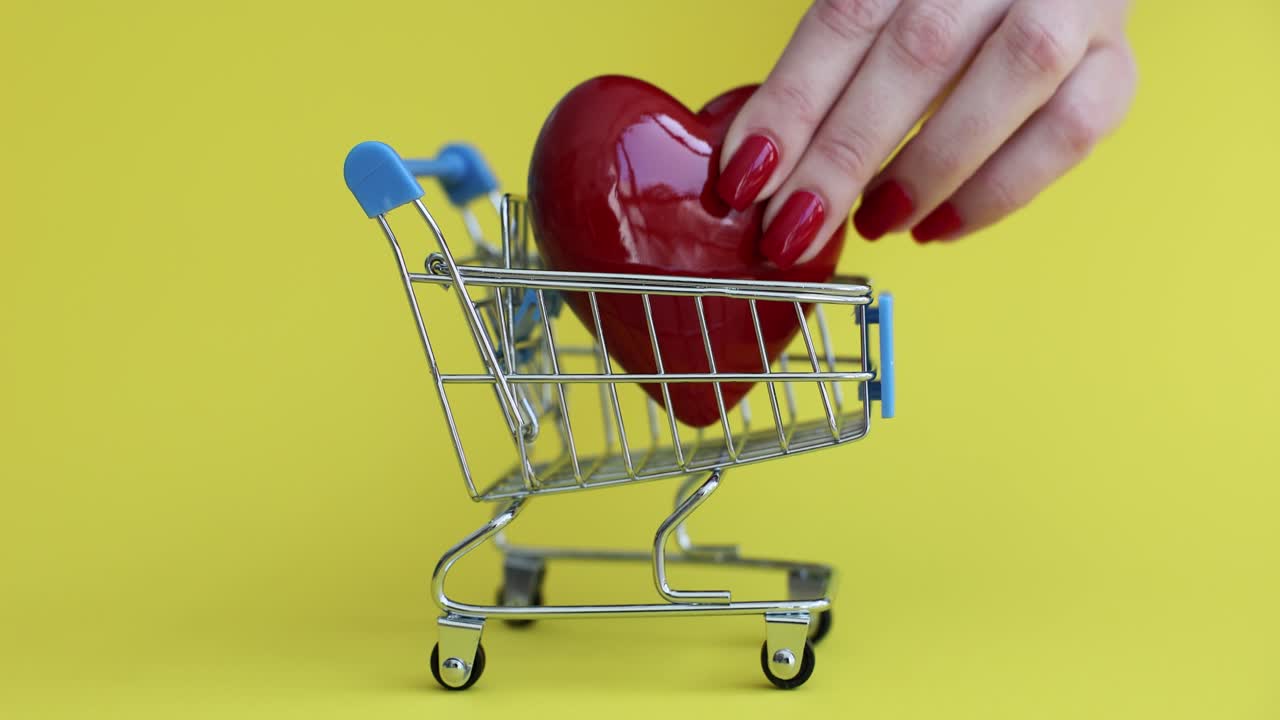 Hand Placing Red Heart into Miniature Shopping Cart on Yellow Background