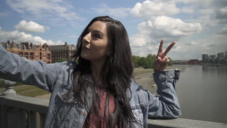 brunette latina tourist taking a selfie, posing doing victory sign with her hand, while standing on the railing of a bridge in london