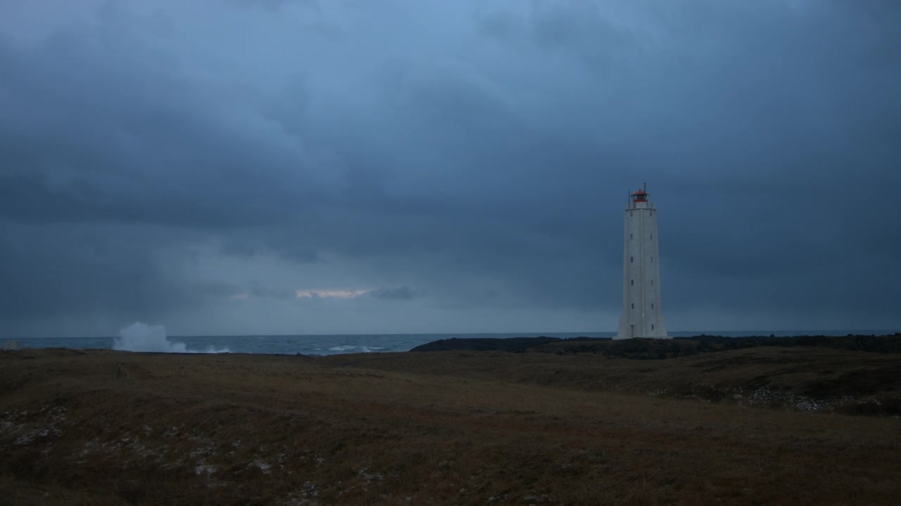 Onde che si infrangono sulla riva in una giornata cupa. Ripresa stabile con un faro in primo piano.