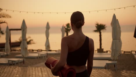 a girl in a black summer sports uniform with a mat in her hands goes to the beach to do morning exercises and yoga. golden colors of the morning beach at sunrise