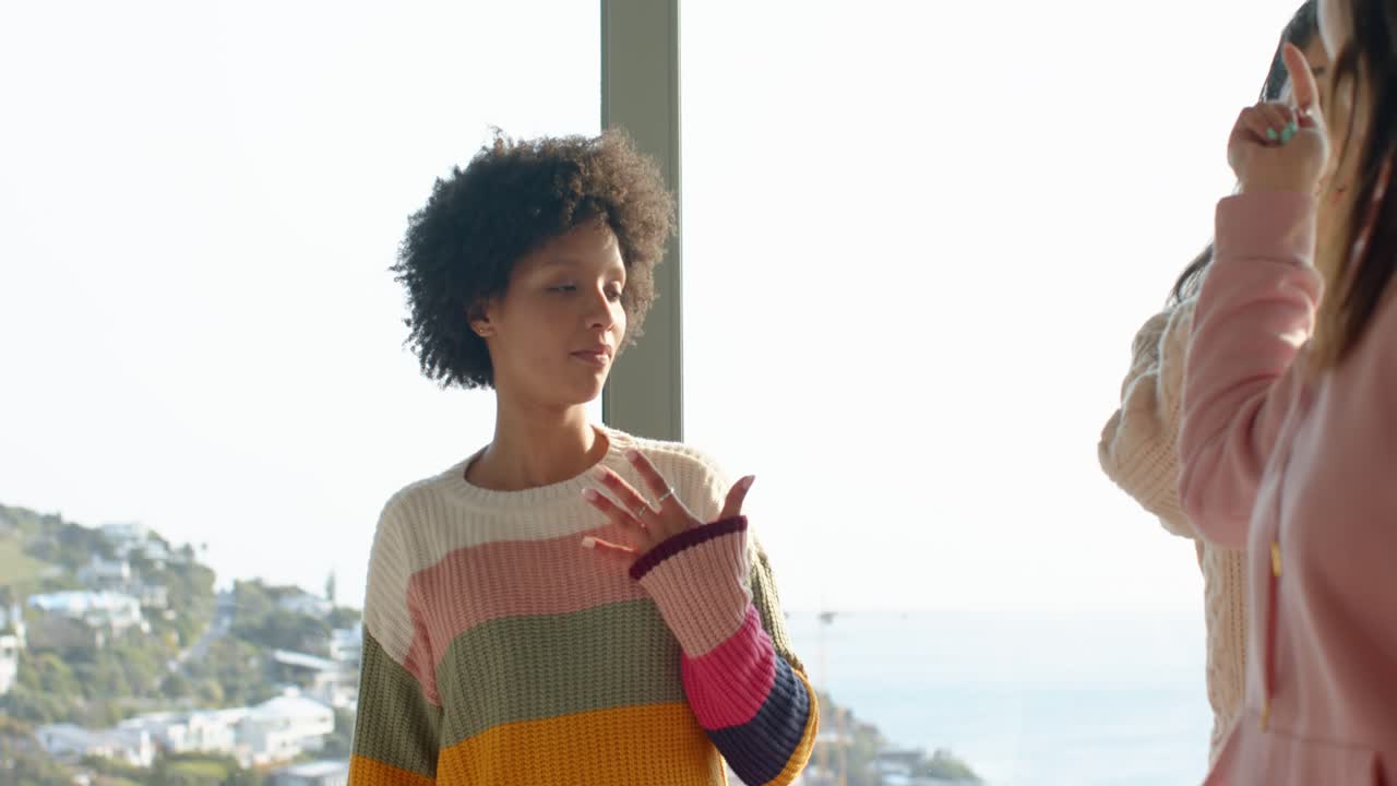 Diverse female friends copying arm choreography being led by pink-hoodie near seaside window
