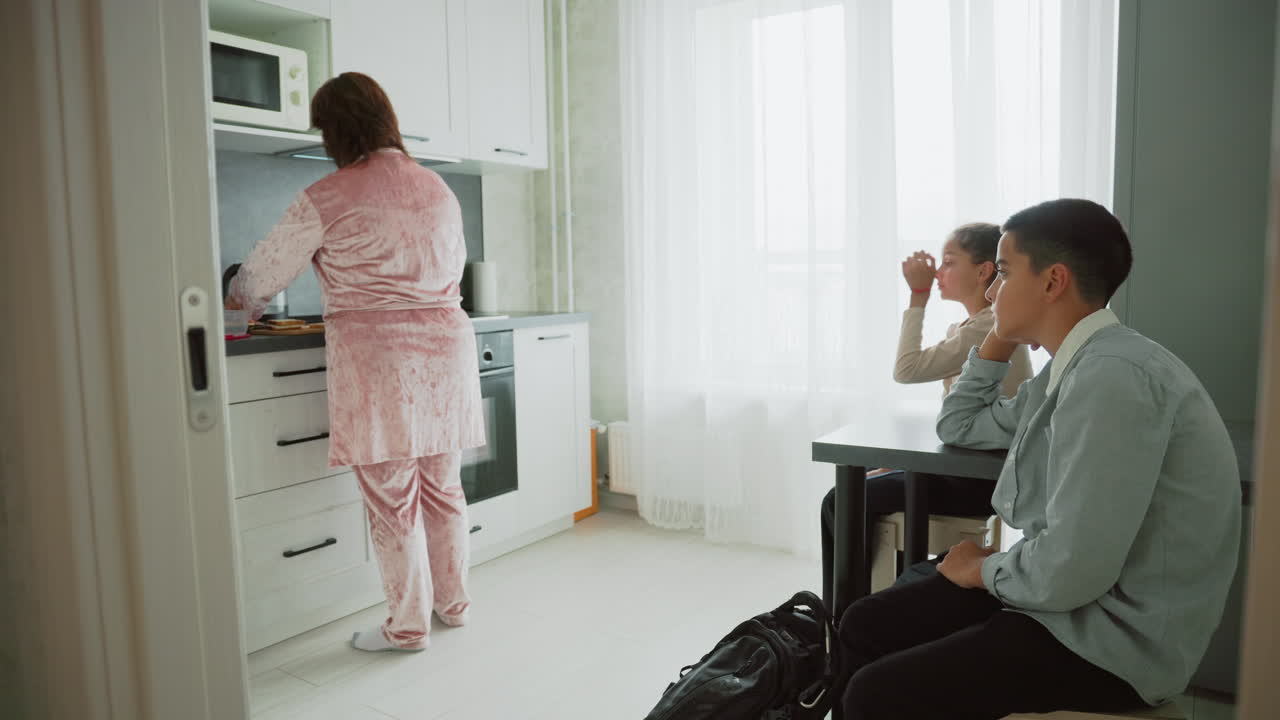 mother wearing pink pajamas cooking breakfast on kitchen countertop while two children in school uniforms sit at table waiting impatiently with backpacks near bright window morning routine scene