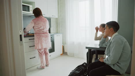 mother wearing pink pajamas cooking breakfast on kitchen countertop while two children in school uniforms sit at table waiting impatiently with backpacks near bright window morning routine scene