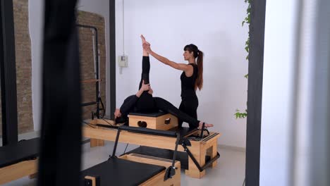 Mujer-Estirando-Las-Piernas-En-Pilates-Reformer-Con-La-Ayuda-De-Un-Entrenador-Personal