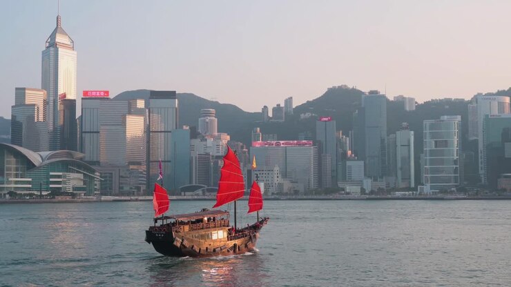 blick auf die uferpromenade des hafens von victoria, während eine hölzerne dschunke mit roten segeln, die alten chinesischen segelschiffen nachempfunden ist und heute als touristenattraktion genutzt wird, vor der skyline der insel hongkong segelt