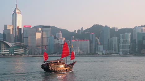 View-of-Victoria-Harbour-waterfront-as-a-wooden-red-sailed-junk-boat-based-on-ancient-Chinese-sailing-ships,-now-used-as-a-touristic-attraction,-sails-in-front-of-the-Hong-Kong-Island-skyline