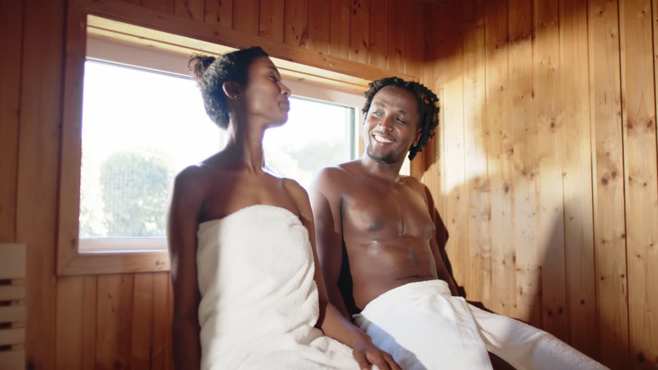 Pareja afroamericana compartiendo sonrisas después de que la mujer se gira hacia el hombre en un banco en la sauna, con toallas.