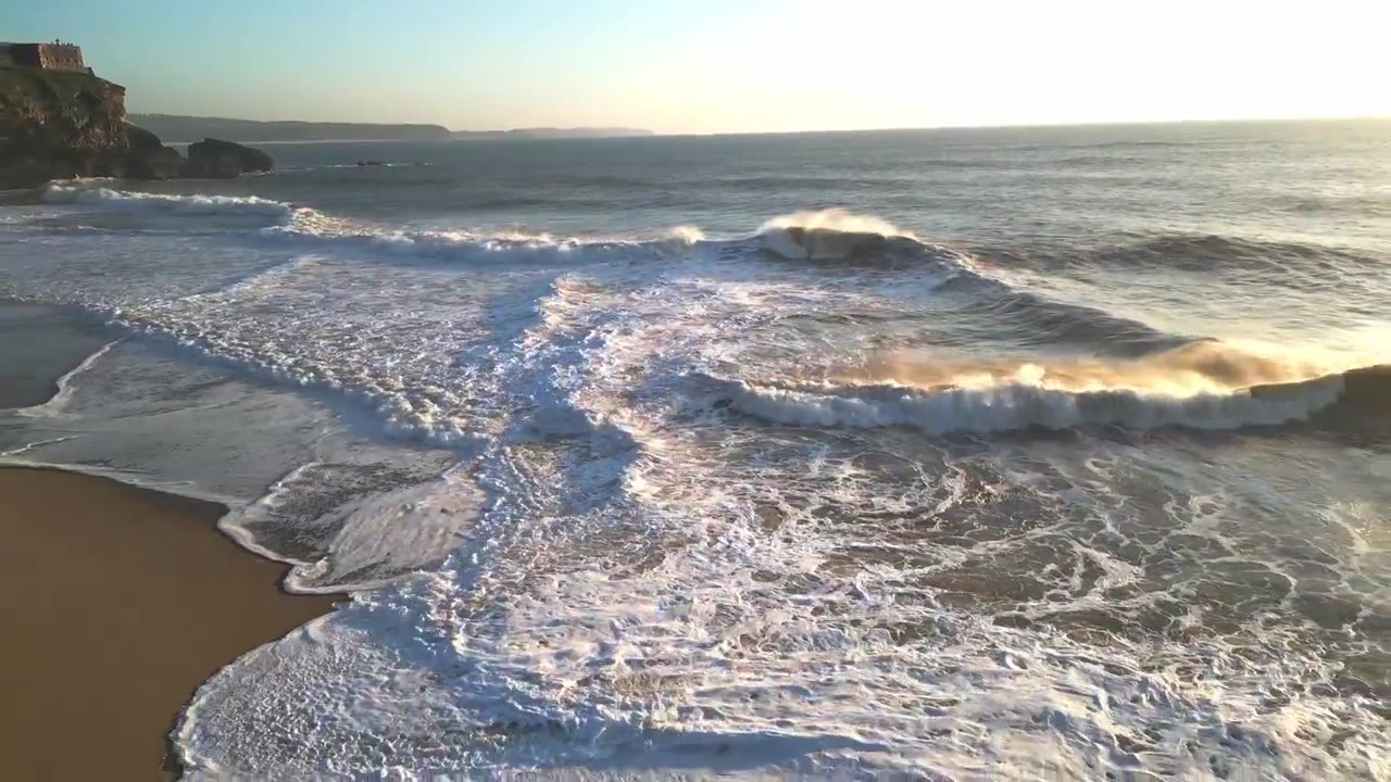 Breathtaking slow motion aerial of the Portuguese coast at sunset. Golden light reflects on crashing waves and white sea foam near rocky cliffs. Cinematic travel footage for atmospheric projects