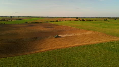 drone shot of a green john deere tractor preparing the field for plantation with a cloud of dust behind tractor on a sunny summer day, following shot