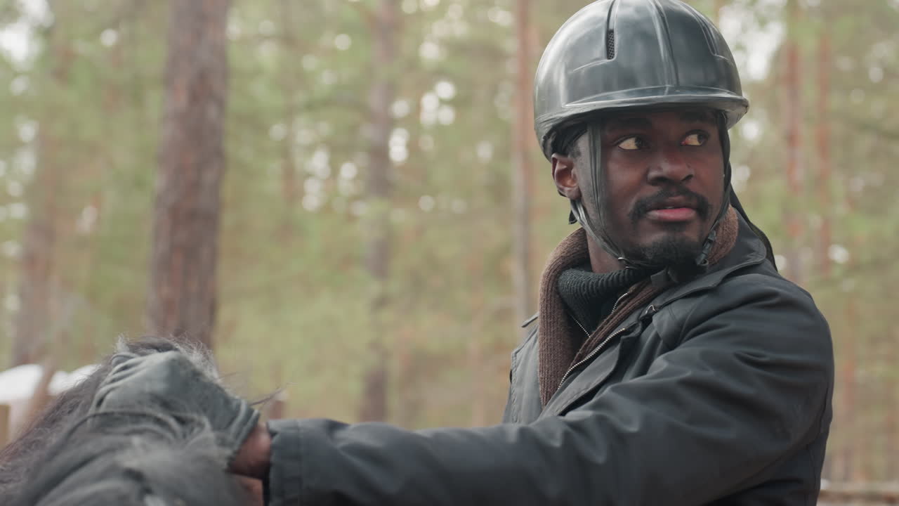 guía con rastro negro cabalgando por un bosque de pinos, con las manos firmes en las riendas y la mirada fija al frente, chaqueta de varias capas y casco contra la fría luz del bosque, cabeza de caballo visible en primer plano, entorno natural al aire libre