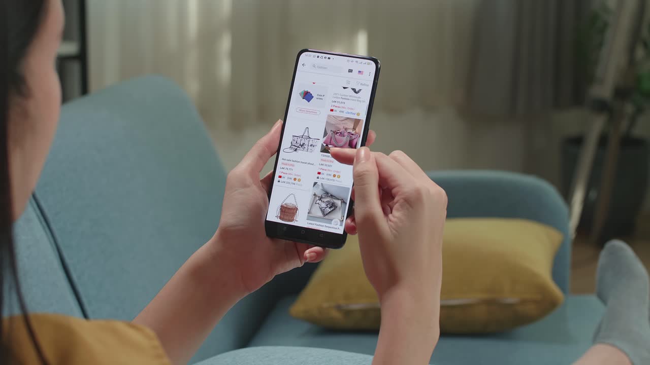 Woman Sitting On Couch In Living Room Using Smartphone Buys In Internet Shop. Female Looks At Goods In Online Store Bag. Buy Fashion Directly On Smartphone