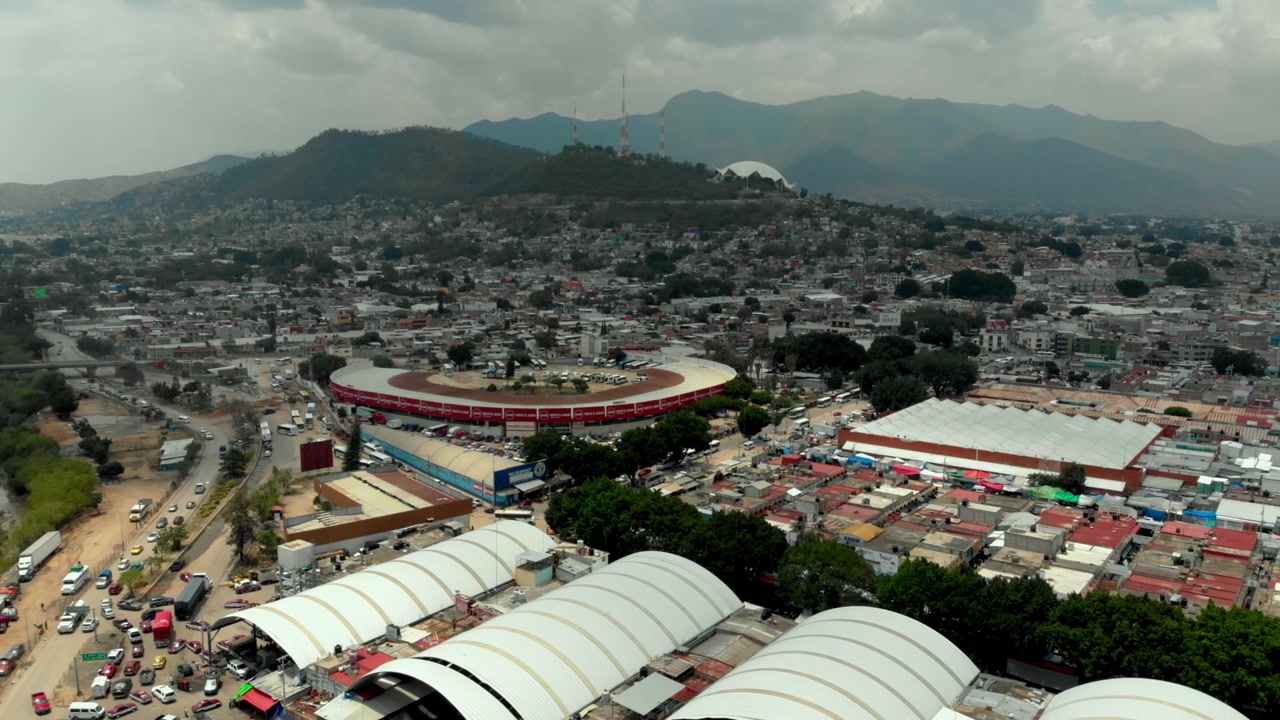 Ripresa aerea con drone di una città densamente popolata con un grande stadio, tetti di mercati, strade trafficate e uno sfondo montuoso sotto la luce del giorno parzialmente nuvolosa, con edifici circostanti.