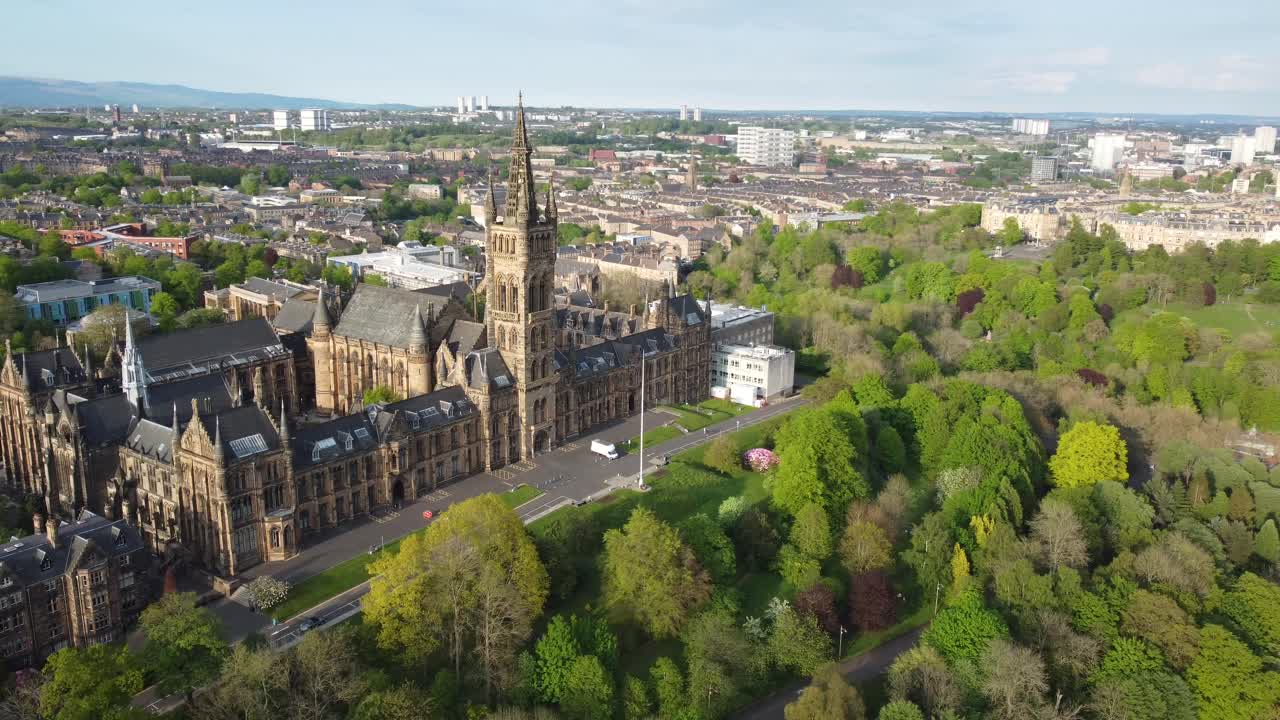 Aerial drone view captures University of Glasgow in Scotland, showing iconic gothic architecture surrounded by lush green park and cityscape under clear blue sky in beautiful daylight