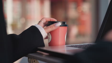 close-up of lady's hand gently turning coffee cup on table next to laptop with blurred background featuring chair, focusing on manicured nails, rings, and cozy workspace setting outdoors