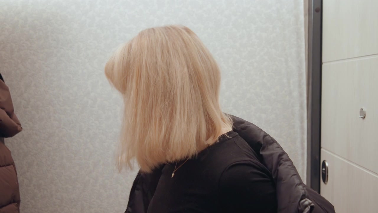 Mujer blanca en el umbral de una puerta con chaqueta, melena rubia de perfil girando hacia la salida, fondo neutro suave, momento de tranquila despedida y anticipación, sutil melancolía y quietud, apartamento urbano
