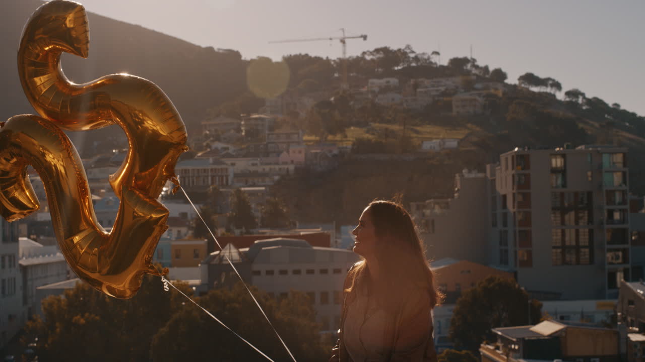 giovane donna felice che celebra la festa di compleanno con palloncini d'oro che galleggiano sul tetto soleggiato al tramonto con lo skyline della città sullo sfondo