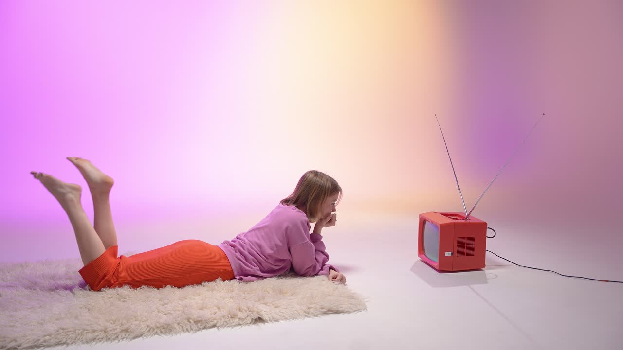 A young woman in bright casual clothes lies on a soft rug, watching a vintage red TV. The pastel lighting enhances the cozy retro mood, blending nostalgia with soft, dreamy studio tones