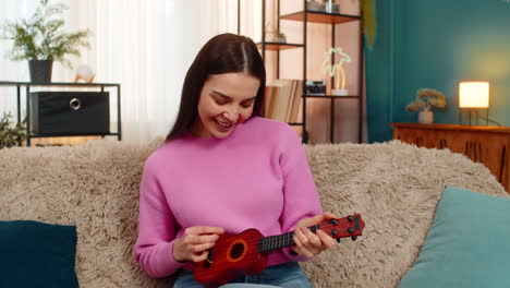 young woman sitting on sofa happily playing toy ukulele instrument smiling joyful cheerful enjoying