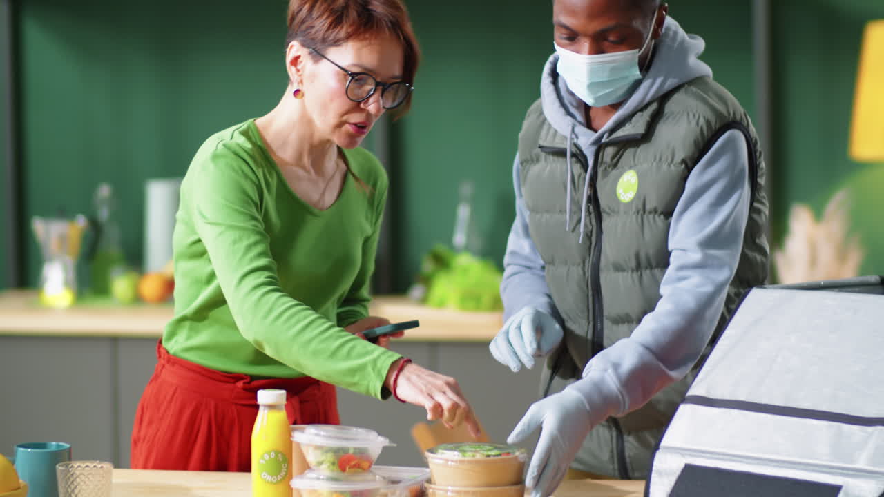 mensajero negro con máscara y guantes descargando cajas de comida para una cliente femenina