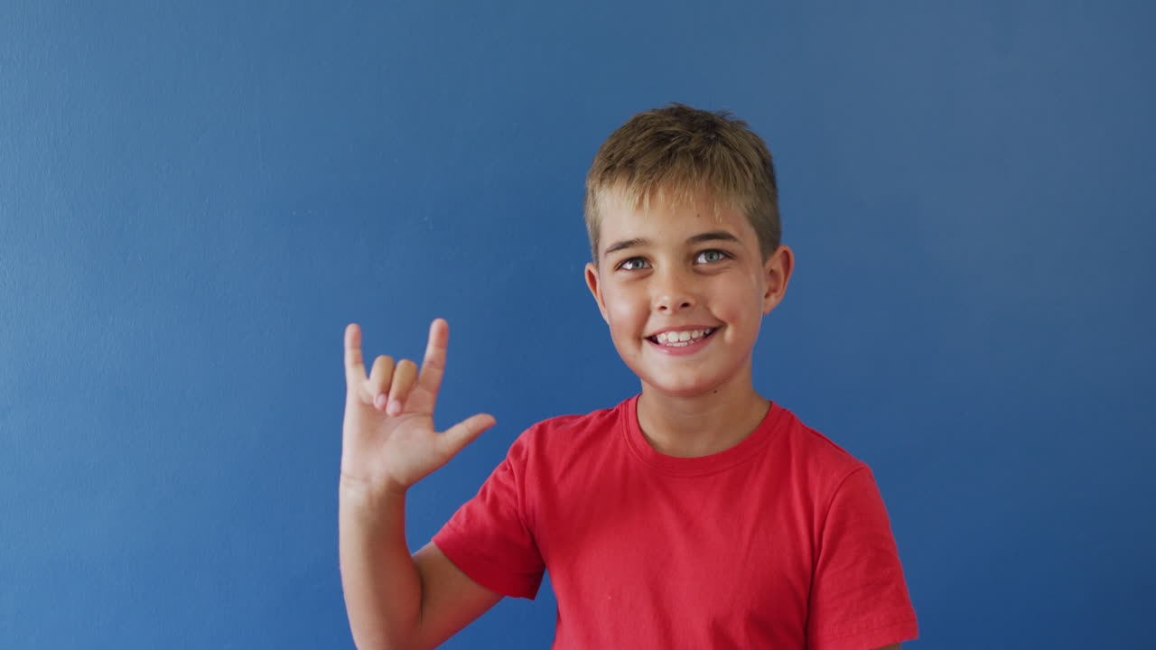 niño caucásico feliz usando lenguaje de señas y sonriendo sobre fondo azul, cámara lenta