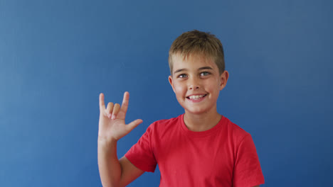 niño caucásico feliz usando lenguaje de señas y sonriendo sobre fondo azul, cámara lenta