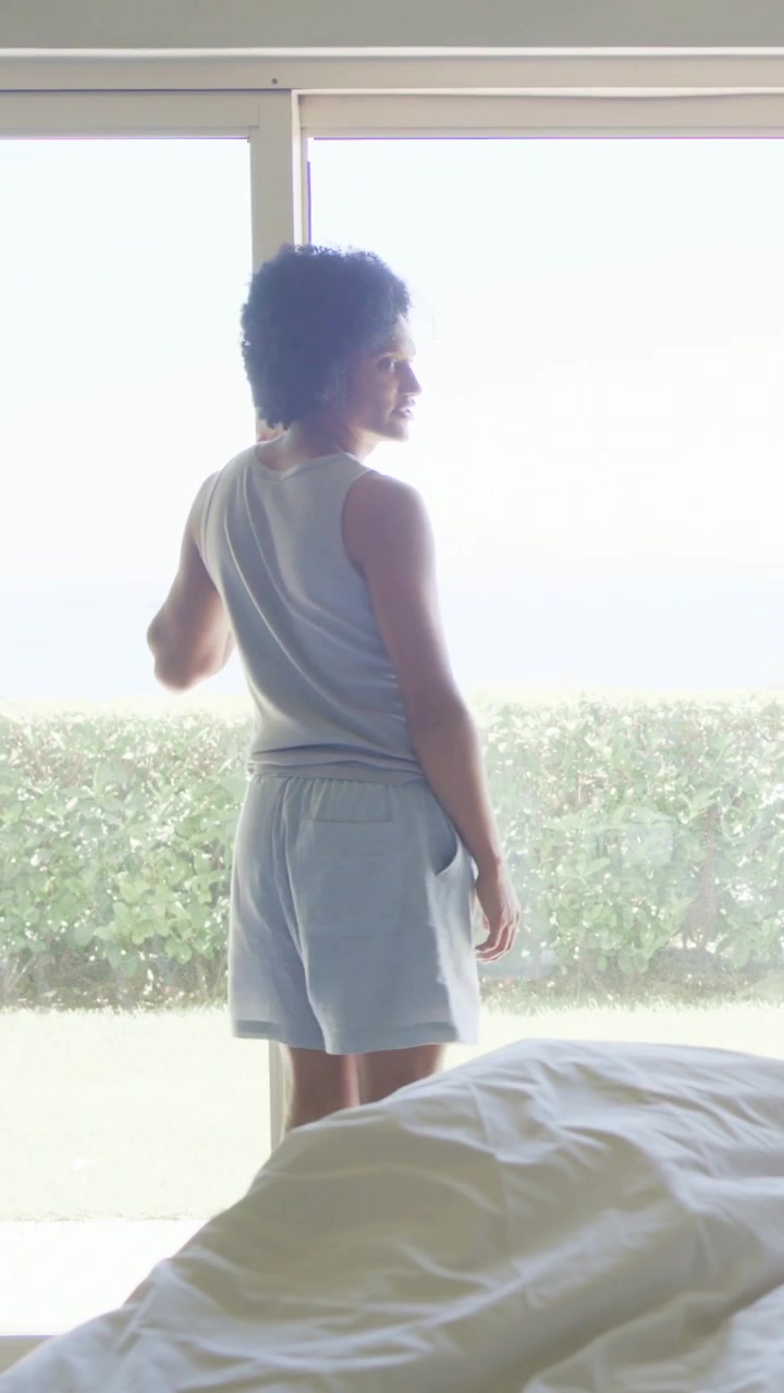 Video verticale: Uomo in canottiera reagisce al sole, apre le tende della porta scorrevole e guarda fuori dalla camera da letto.