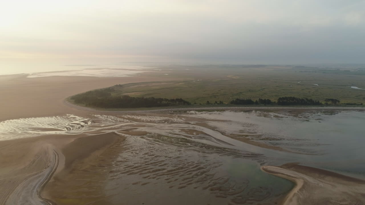 cinemática que establece un disparo de un dron durante la marea baja hacia una playa de arena con árboles y marismas detrás al amanecer en la costa este del reino unido de north norfolk