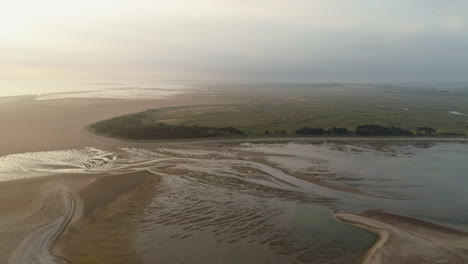 cinematic establishing drone shot at low tide towards sandy beach with trees and salt marsh behind at sunrise in north norfolk uk east coast