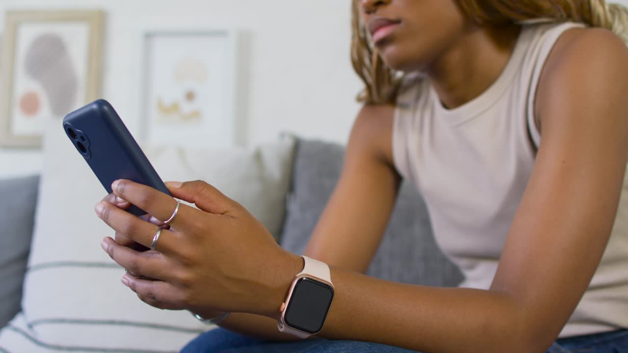 African American woman holding smartphone, tapping and reading message on sofa, smartwatch visible